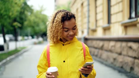 Lächelndes-Afroamerikanisches-Mädchen-nutzt-Smartphone-SMS-Freunde-und-halten-zum-Mitnehmen-Kaffee-zu-Fuß-in-die-Stadt-allein.-Moderne-Technik,-Kommunikation-und-Getränke-Konzept.