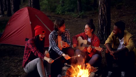 Mujer-bonita-es-tocar-la-guitarra-mientras-sus-amigos-cantando-canciones-divertidas-y-risa-melcocha-cocción-en-fuego-y-palmas-de-las-manos.-Concepto-de-música,-naturaleza-y-diversión.