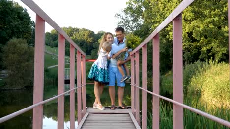 Familia-feliz:-Papá,-mamá-y-pequeño-hijo-parado,-abrazándose-y-besándose-en-el-puente-en-el-parque-de-la-laguna.-El-hijo-se-encuentra-en-las-manos-del-Papa.