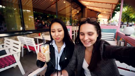 Point-of-view-shot-of-attractive-ladies-making-online-video-call-holding-device-with-camera-and-talking-clinking-cocktail-glasses-and-showing-shopping-bags-looking-at-camera.