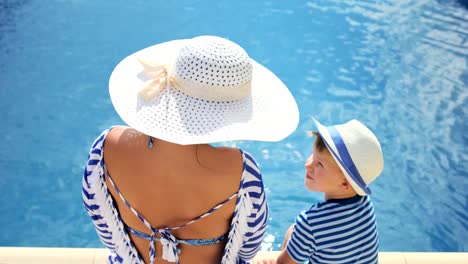 Mujer-con-sombrero-blanco-con-arco-y-niño-sentados-en-el-borde-de-la-piscina-juntos