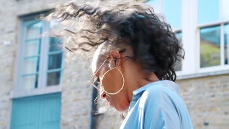 Fashionable-young-black-woman-wearing-blue-dress-and-sunglasses-laughing-to-camera-in-the-street,-backlit,-low-angle,-close-up