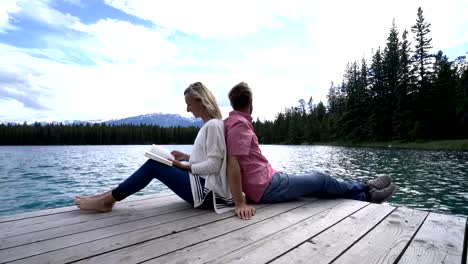 Couple-relaxing-on-wooden-lake-pier,-sitting-back-to-back,-Canada