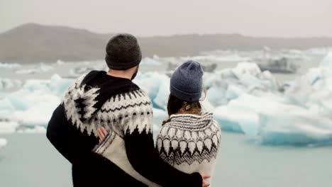 Back-view-of-young-couple-standing-in-ice-lagoon.-Man-kisses-the-woman-and-hugs-her,-looking-on-glaciers-in-Iceland