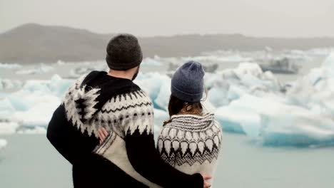 Vista-posterior-de-pie-de-joven-pareja-en-la-laguna-de-hielo.-Hombre-besa-a-la-mujer-y-la-abraza,-en-los-glaciares-en-Islandia