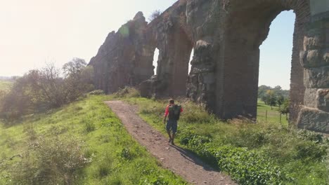 Hombre-joven-mochilero-caminando-por-camino-de-tierra-a-lo-largo-del-antiguo-acueducto-romano-en-naranja-ropa-deportiva-senderismo-dolly-de-drone-de-vista-aérea