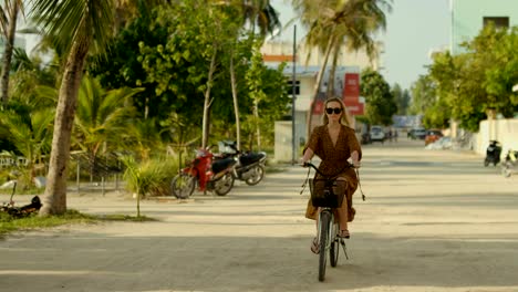 Beautiful-Woman-Wearing-Dress-Rides-Bicycle-on-the-Road-Through-Authentic-Small-Town.-South-Asia-Nature,-Scenic-Forest-Growing-by-the-Road.