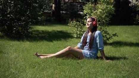 Chica-joven-escuchando-música-con-auriculares-con-smartphone-sentado-sobre-la-hierba-en-el-parque-en-tiempo-soleado.-4K