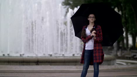 Mädchen-mit-einem-Regenschirm-auf-einem-Hintergrund-von-einem-Brunnen-im-Park-spazieren-in-die-Kamera-schaut