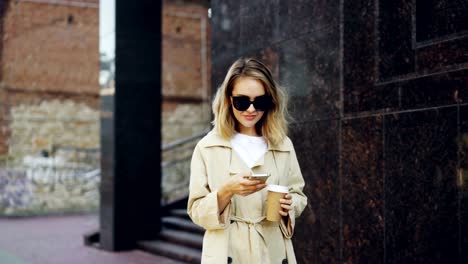 Elegant-young-lady-is-walking-along-street,-using-smart-phone-and-drinking-to-go-coffee-enjoying-walk-in-beautiful-modern-city.-People,-modern-lifestyle-and-takeaway-drinks-concept.