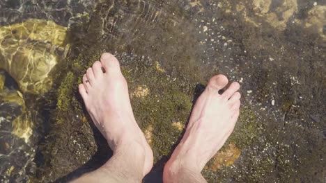 Male-legs-standing-on-stones-in-river-water-point-of-view.-Barefoot-man-in-river