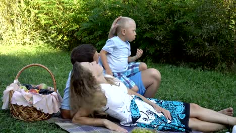 Family-picnic-in-the-Park.-Mom-and-dad-lie-on-the-grass,-and-the-happy-little-son-sits-on-top-of-them-and-eats-grapes.