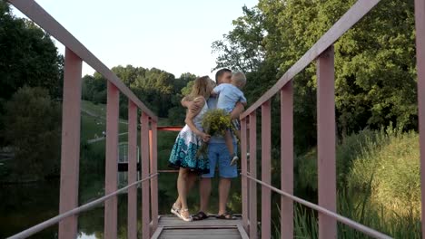 El-feliz-papá,-mamá-e-hijo-pequeño-están-parado-en-el-puente-sobre-el-estanque-en-el-parque-contra-el-paisaje.-El-hijo-se-sienta-en-las-manos-del-Papa,-besos-y-abrazos-le-con-su-madre.