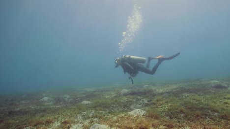 Scuba-diver-floating-on-sea-bottom-and-waving-hand-to-camera,-underwater-view