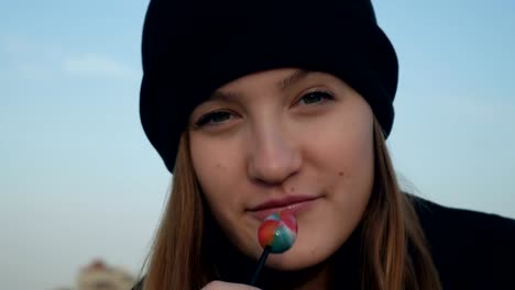 Hübsches-Teenager-Mädchen-in-schwarzem-Hut-essen-Lolipop,-Blick-in-die-Kamera-und-lächelnd,-hautnah-erschossen,-im-Freien