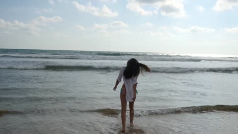 Happy-woman-in-bikini-and-shirt-walking-on-the-beach-near-the-ocean.-Young-beautiful-girl-enjoying-life-and-having-fun-at-sea-shore.-Summer-vacation-or-holiday.-Rear-back-view