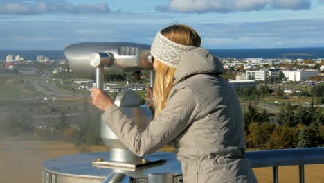 young-tourist-girl-is-looking-in-a-stationary-telescope-on-Reykjavik-in-sunny-day