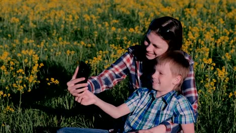 Mutter-und-Sohn-machen-Selfie-am-Telefon-sitzen-auf-dem-Rasen-unter-den-gelben-Blumen.