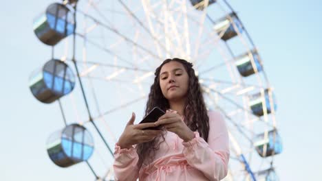 A-girl-with-long-hair-in-a-pink-dress-makes-selfie-using-a-smartphone-standing-near-the-Ferris-wheel.-4K