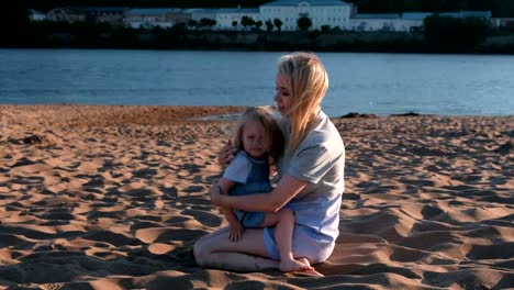 Beautiful-happy-blonde-mom-and-daughter-cuddling-and-speaking-sitting-on-the-beach-at-sunset.