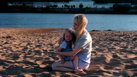 Hermosa-mamá-rubia-feliz-e-hija-abrazando-y-hablando-sentados-en-la-playa-al-atardecer.