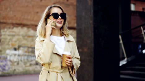 Beautiful-young-woman-with-blond-hair-is-talking-on-mobile-phone-and-drinking-take-away-coffee-walking-in-city-along-street-and-enjoying-drink-and-conversation.