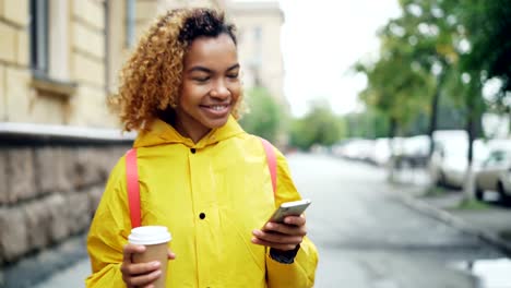 Glückliche-afroamerikanische-Frau-nutzt-Smartphone-Bildschirm-berühren-und-lächelnd,-walking-im-freien-in-schönen-Stadt-mit-Kaffee-zum-Mitnehmen.-Modernen-Lebensstil-und-Kommunikationskonzept.