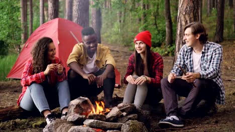 Los-alegres-turistas-hombres-y-mujeres-comparten-historias-sentados-alrededor-de-fogatas-y-riendo-está-calentando-cerca-de-ardiente-llama.-Concepto-de-conversación,-gente-y-diversión.