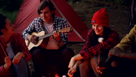 Freunde-auf-Campingplatz-sind-Lieder-singen,-lachen-und-Essen-auf-Feuer-Kochen,-während-glücklich-lächelnden-Mann-der-Gitarre-spielt.-Camping,-Musik-und-Freundschaft-Konzept.