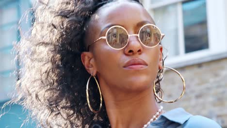 Fashionable-young-black-woman-wearing-blue-dress-and-sunglasses-standing-on-the-street,-backlit,-head-and-shoulders-low-angle