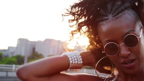 Close-up-of-young-black-woman-wearing-sunglasses-plying-with-her-hair,-looking-to-camera-on-a-London-street-at-the-golden-hour