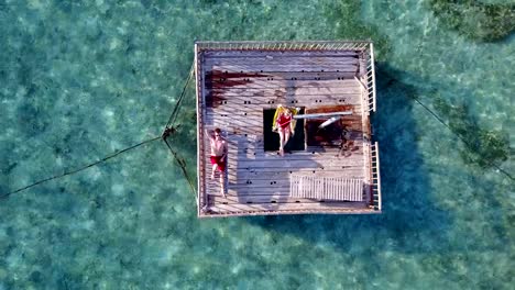 v03858-Aerial-flying-drone-view-of-Maldives-white-sandy-beach-2-people-young-couple-man-woman-relaxing-on-sunny-tropical-paradise-island-with-aqua-blue-sky-sea-water-ocean-4k-floating-pontoon-jetty-sunbathing-together