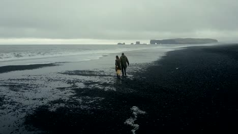 Aerial-view-of-young-stylish-couple-walking-on-the-black-volcanic-beach-in-Iceland.-Man-and-woman-running-from-the-wave