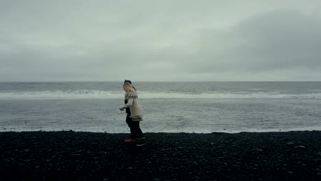 Copter-flying-near-the-young-couple-in-icelandic-sweater.-Man-and-woman-running-on-the-black-volcanic-beach