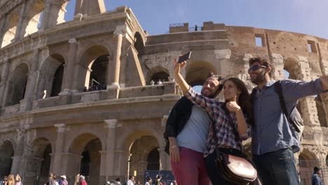 Three-young-friends-tourists-standing-in-front-of-colosseum-in-rome-taking-selfies-with-smartphone-with-backpacks-sunglasses-happy-beautiful-girl-long-hair-slow-motion