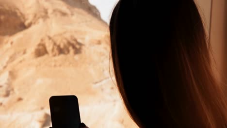 Girl-using-smartphone-in-aerial-railway-cabin.-Woman-takes-phone-photos-in-sunny-desert-ropeway-car.-Masada,-Israel.-4K