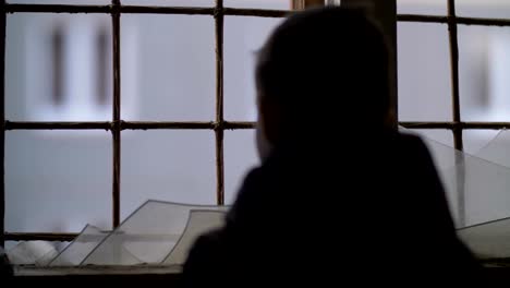 back-of-refugee-boy-stands-alone-near-a-broken-window,-looking-out-the-window,-ban-on-a-good-life