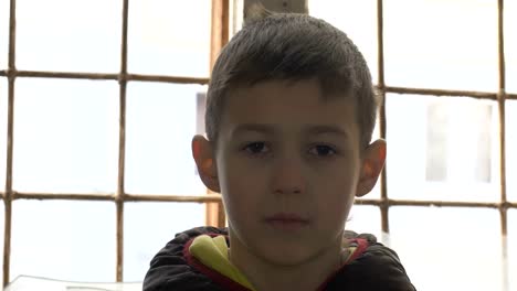 portrait-emaciated-refugee-boy-stands-alone-near-a-broken-window,-looking-into-the-camera