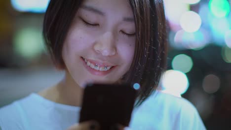 Portrait-of-the-Attractive-Japanese-Girl-Wearing-Casual-Clothes-Uses-Smartphone.-In-the-Background-Big-City-Advertising-Billboards-Lights-Glow-in-the-Night.