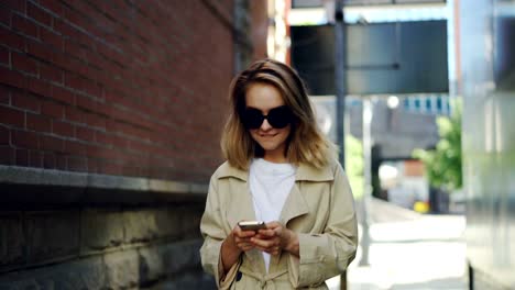 Pretty-blond-girl-is-using-modern-smartphone-touching-screen-walking-in-city-and-smiling.-Young-woman-is-wearing-fashionable-clothing-and-trendy-sun-glasses.