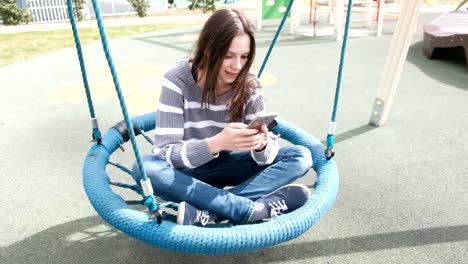 Beautiful-brunette-woman-is-swinging-on-empty-webbed-swing,-loking-at-social-media-on-mobile-phone.