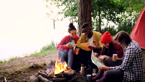 Cinemagraph-loop---group-of-young-men-and-women-are-looking-at-paper-maps-sitting-around-fire-in-forest-with-drinks.-Map-is-moving,-tent-and-trees-are-visible.