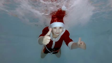 Un-niño-vestido-de-Santa-Claus-nada-bajo-el-agua-en-los-chorros-de-agua-en-la-piscina-con-su-sonrisa-abierta,-ojos,-mira-a-la-cámara-y-demuestra-sus-dedos-para-arriba.