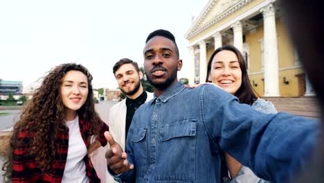 Point-of-view-shot-of-happy-friends-tourists-making-online-video-call-holding-device,-looking-at-camera,-laughing-and-talking-about-sights-in-beautiful-city-around-them.