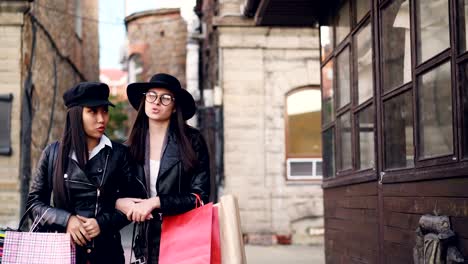 Happy-girls-in-trendy-clothing-are-walking-arm-in-arm-along-street-carrying-shopping-bags-and-talking-socializing-on-autumn-day.-People,-conversation-and-fashion-concept.