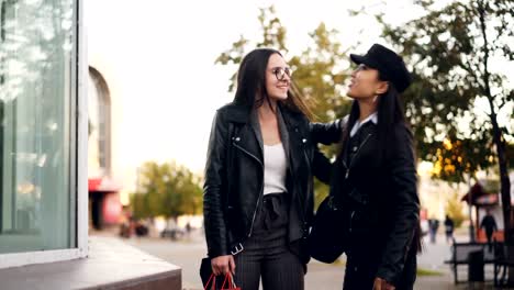 Attractive-girl-is-choosing-clothes-from-shop-window-standing-outdoors-then-meeting-her-friend,-young-women-are-hugging,-discussing-things-in-paper-bags-and-leaving.