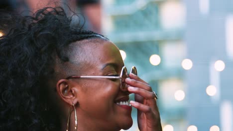 Moda-joven-mujer-de-negra-en-gafas-de-sol-mirando-a-su-alrededor,-un-disparo-de-cabeza,-luces-bokeh-en-fondo