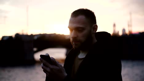 Retrato-de-joven-barbudo-gastar-tiempo-en-terraplén-hermoso,-usando-el-teléfono-inteligente-en-la-noche,-el-atardecer