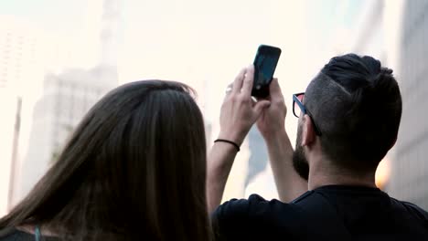 Young-beautiful-couple-walking-in-downtown-of-Chicago,-America-and-taking-photo-of-skyscraper-on-smartphone