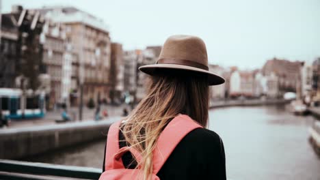 Dame-mit-rotem-Rucksack-steht-auf-Flussbrücke.-Mädchen-im-stylischen-Hut-mit-langen-Haaren-und-einem-Telefon-warten.-4K-Rückansicht